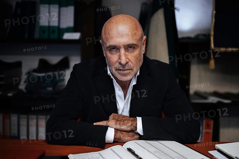 Portrait of a confident businessman seated at a desk, surrounded by office files. The scene conveys focus and determination in a professional corporate setting