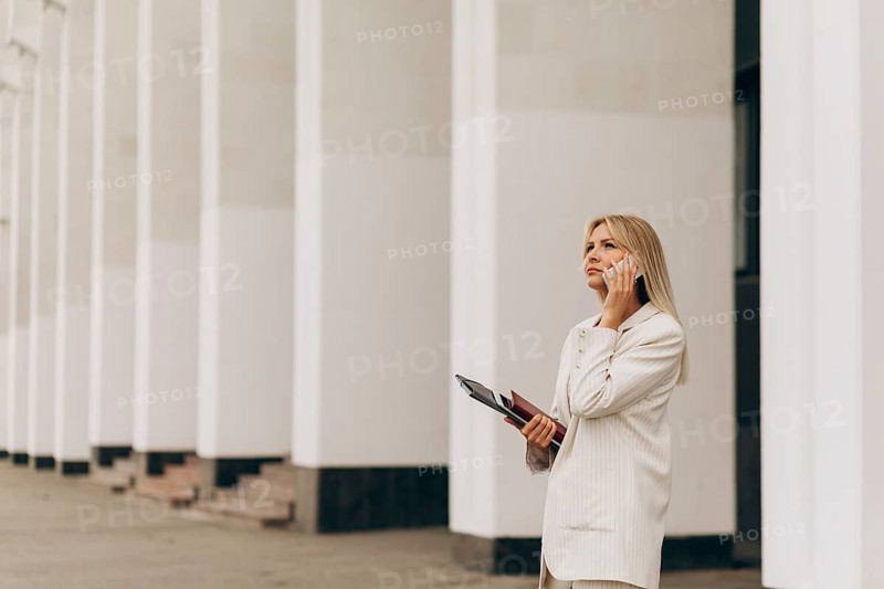 A businesswoman in a beige suit talks on her phone while standing outside an office building with white columns. She holds a folder, looking focused and professional