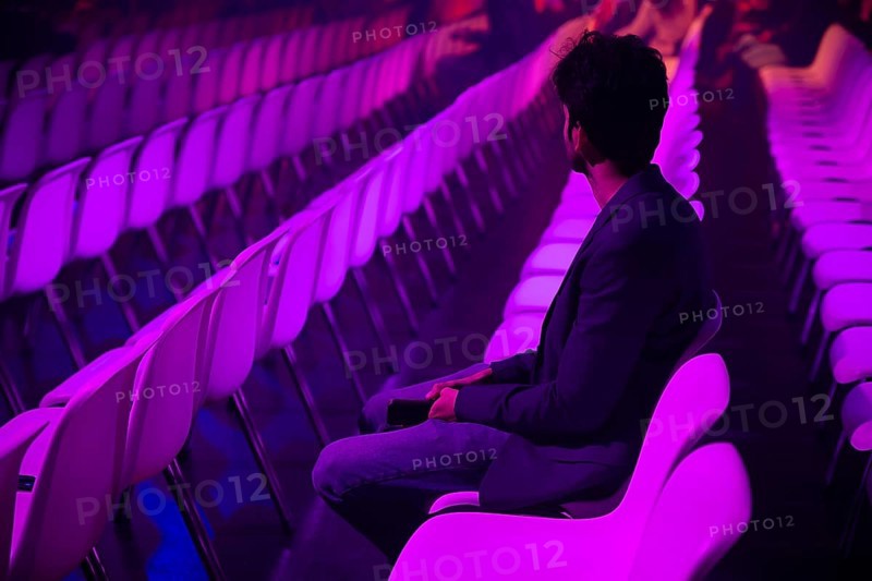 A indian man on a white chair in a spacious, purple lit auditorium at a conference of AI in Amsterdam. The atmosphere is serene with rows of empty seats