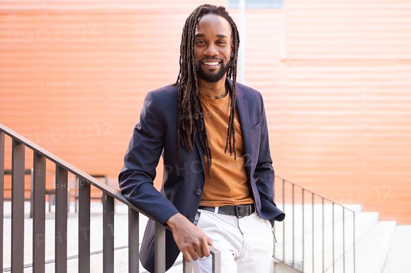 Cuban business man with long hair wearing a blazer and casual attire, leaning against a staircase railing outdoors, smiling warmly. Stylish urban setting with orange wall