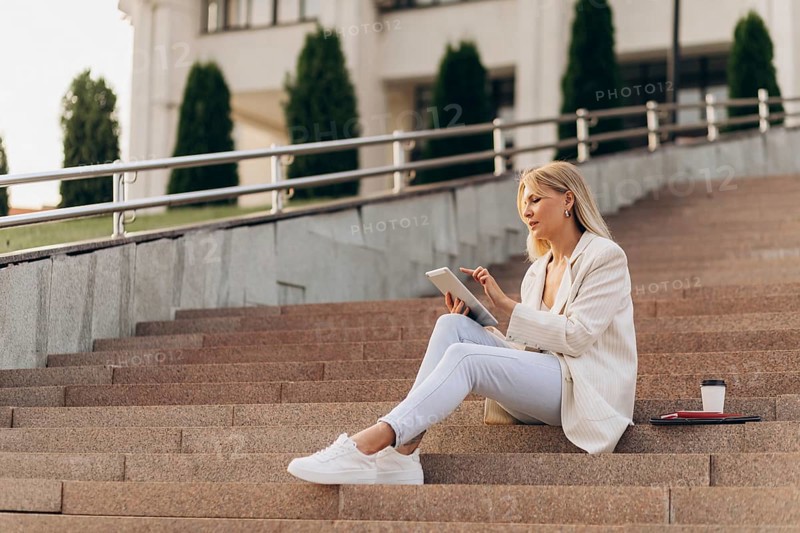A businesswoman in casual attire sits on outdoor steps with a digital tablet, concentrating on her work. A takeaway coffee cup and papers lie beside her, emphasizing a modern urban lifestyle
