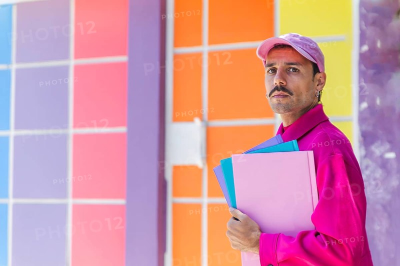 A young Spanish Caucasian man with a mustache, dressed in vibrant pink clothing, holding folders against a colorful backdrop. He embodies a cheerful urban lifestyle and casual fashion, expressing individuality and diversity