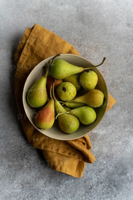 A top view of a ceramic bowl filled with fresh wild pears, set on a rustic surface with a brown cloth underneath. The image captures the natural colors and textures