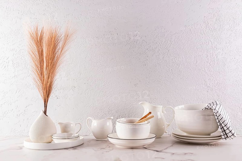 A minimalist interior design featuring elegant white tableware and a decorative vase with dried grass, arranged tastefully on a marble surface against a textured white wall