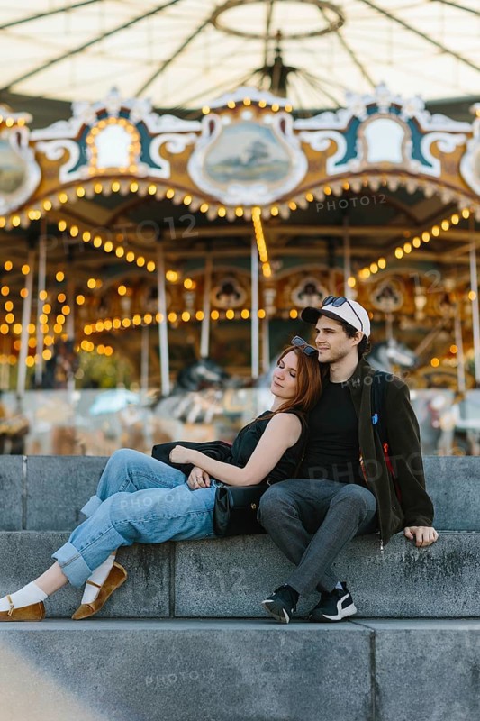 A couple sits on stone steps, the woman leaning on the man, in front of a brightly lit carousel in Brooklyn Bridge Park. They exude a sense of relaxation and enjoyment