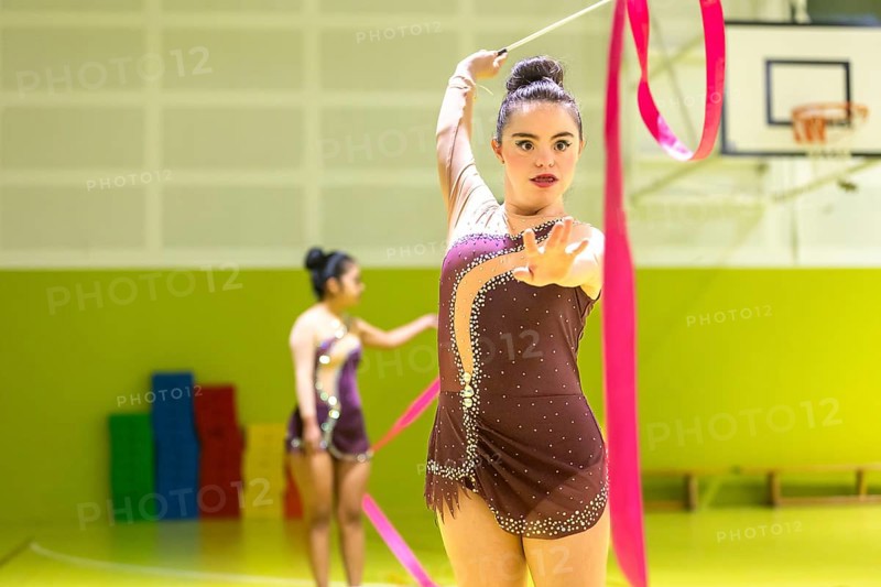 Young disabled rhythmic gymnast in elegant attire performs with a pink ribbon inside a sports hall. Another gymnast practices in the background. Dynamic movement and focus