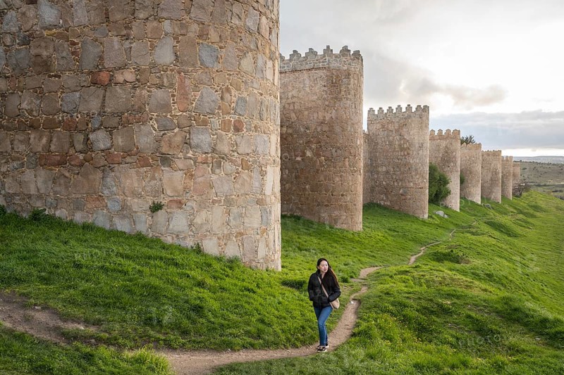 An Asian female tourist strolls along Avila's ancient medieval walls. The scene captures cultural exploration and architectural beauty