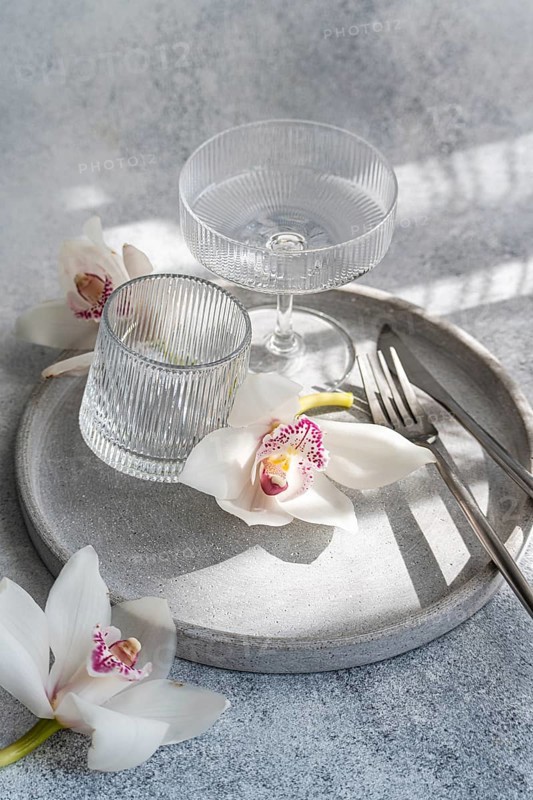 A refined dining setup featuring ribbed glassware, delicate orchids, and sleek cutlery on a circular concrete tray, creating a sophisticated and serene atmosphere