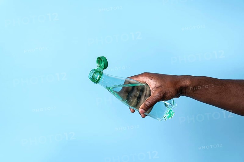 A close-up image of a hand holding a transparent water bottle with a green cap, set against a smooth blue background. The scene portrays a simple, refreshing theme