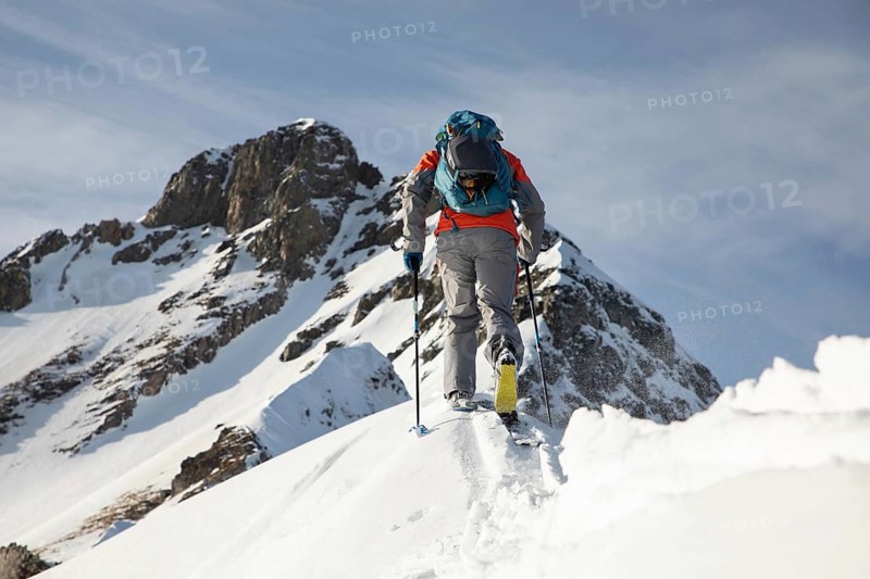 A skier in vibrant gear ascends a snowy peak, showcasing the thrill and challenge of ski touring amidst breathtaking mountain scenery under a clear blue sky