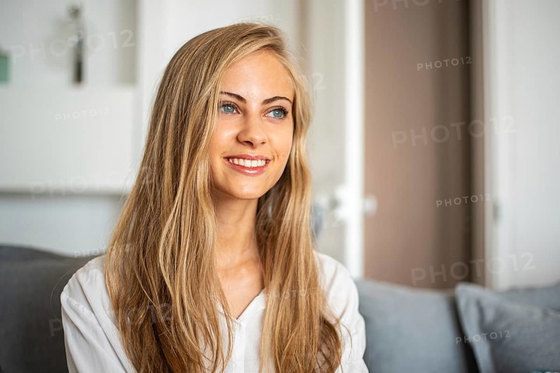 Portrait of a smiling woman with long blonde hair sitting in a bright, cozy room. She looks relaxed and content, capturing a moment of genuine happiness and warmth