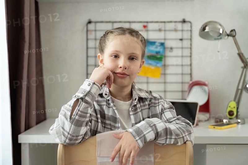 A young girl with braided hair sits on a chair in front of a white desk at home. She looks confidently at the camera in a calm and tidy room with creative touches