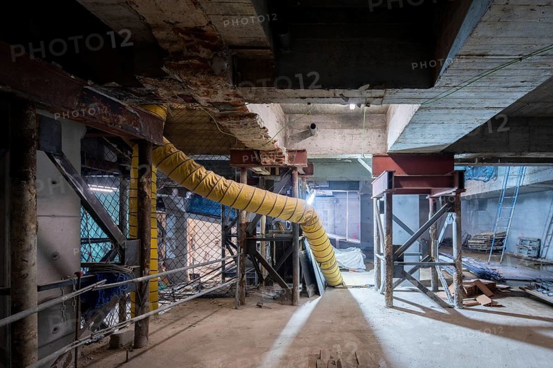 A construction site undergoing structural reinforcement features steel supports, exposed concrete beams, and a large ventilation duct. The dimly lit underground space highlights the ongoing renovation work