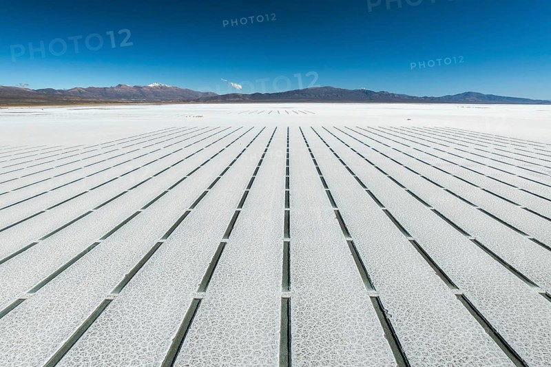 Aerial view of the geometric grid pattern created by salt extraction lines in Salinas Grandes, Salta, Argentina, under a vibrant blue sky with distant mountain ranges in the background