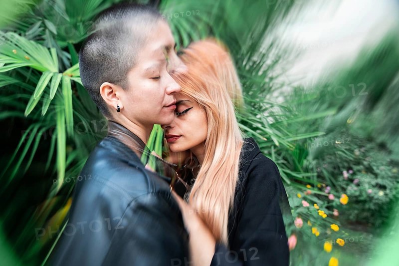 A lesbian couple shares a tender, intimate moment surrounded by vibrant greenery, symbolizing love and connection. The lush foliage adds a serene backdrop to their embrace