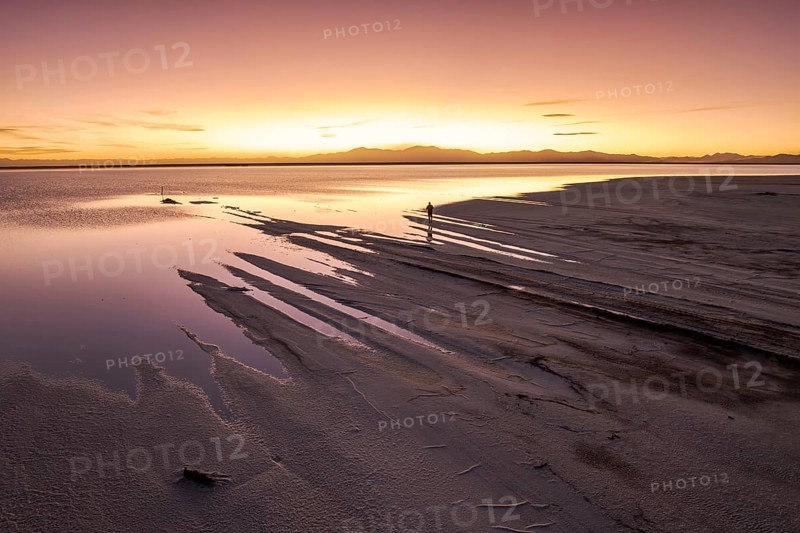 A solo traveler at a mesmerizing sunset in Salinas Grandes, Salta, Argentina, where the vast salt flats meet a golden horizon, creating stunning reflections and a tranquil atmosphere