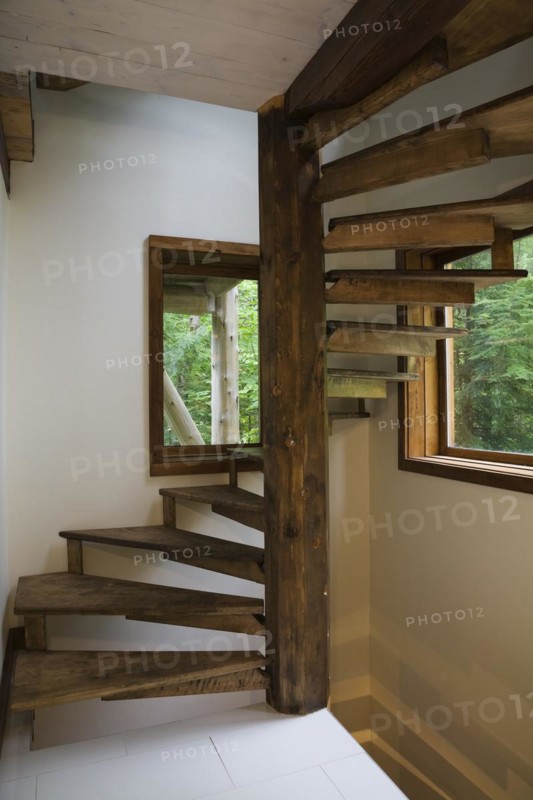 Brown stained cherry wood spiraling staircase with thin flat wooden steps attached to pine wood post inside Scandinavian hybrid style log cabin home, Quebec, Canada