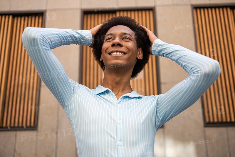 A joyful non-binary individual confidently poses outdoors in a vibrant urban setting, showcasing a bright smile and relaxed posture against a modern architectural backdrop