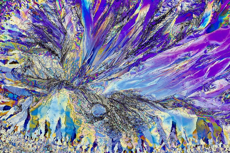 This striking image captures the vibrant and intricate patterns of microcrystals under a microscope. The colors range from deep blues and purples to radiant yellows and pinks, creating a dynamic visual effect