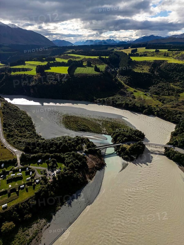 Aerial view captures the diverse landscape of Canterbury, New Zealand, highlighting winding rivers, lush farmlands, a bridge, and distant mountains under a cloudy sky