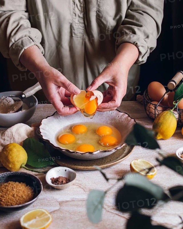A person cracks eggs into a bowl on a table surrounded by lemons, brown sugar, and spices, capturing a rustic kitchen scene filled with fresh ingredients