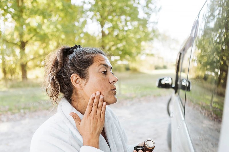 A woman enjoys her skincare routine while experiencing the freedom of van life in autumn. Captured in New Zealand, the serene moment highlights her connection to nature