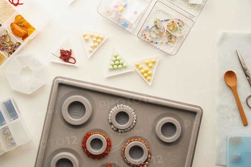 Top view of a creative jewelry making setup in a studio with tools like scissors, a wooden spoon, bead sorting trays, and a bracelet mandrel highlighting handmade craftsmanship