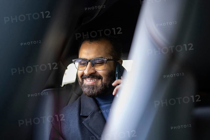 A smiling Indian man dressed in a winter coat and sweater, talking on a mobile phone while sitting in a car, looking away from the camera