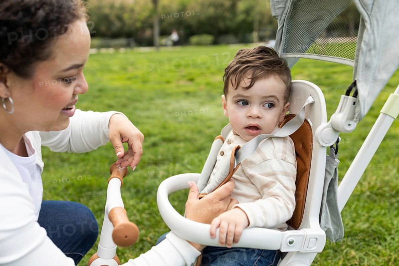 A loving mother bends down to interact with her baby sitting in a stroller. They are enjoying a sunny day at the park, surrounded by lush green grass and peaceful scenery