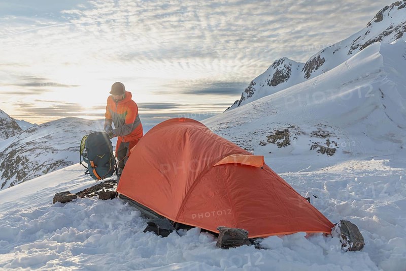 A ski tourer sets up near a bright orange tent amidst snowy mountains at sunrise, capturing the spirit of adventure and outdoor exploration in a stunning winter landscape