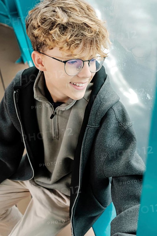 A curly-haired teenager with glasses sits by a window, smiling as he enjoys a ski resort in the Pyrenees. His relaxed posture and comfortable clothing create a moment of serene reflection