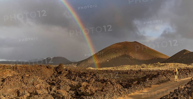 A vibrant rainbow arcs over the rugged volcanic terrain of Timanfaya National Park in Lanzarote, Canary Islands, creating a striking contrast against the stormy sky