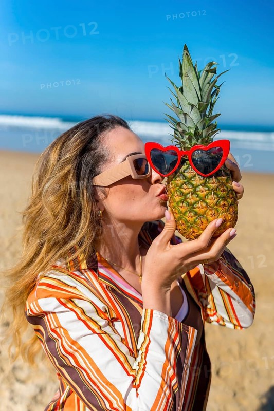 Woman playing with a pineapple wearing red heart sunglasses, standing on a sandy beach by the ocean, embodying summer fun and exotic vacation vibes under a clear blue sky