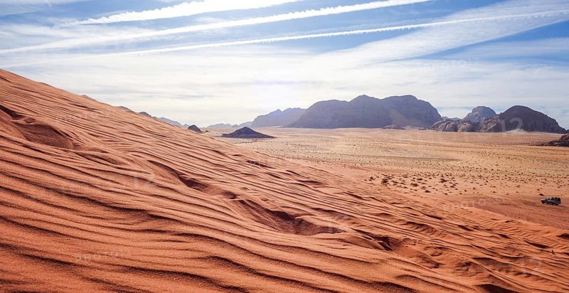 Captivating view over the red sand dunes in Wadi Rum Desert, Jordan, under a clear sky, with distant mountains and a lone vehicle visible
