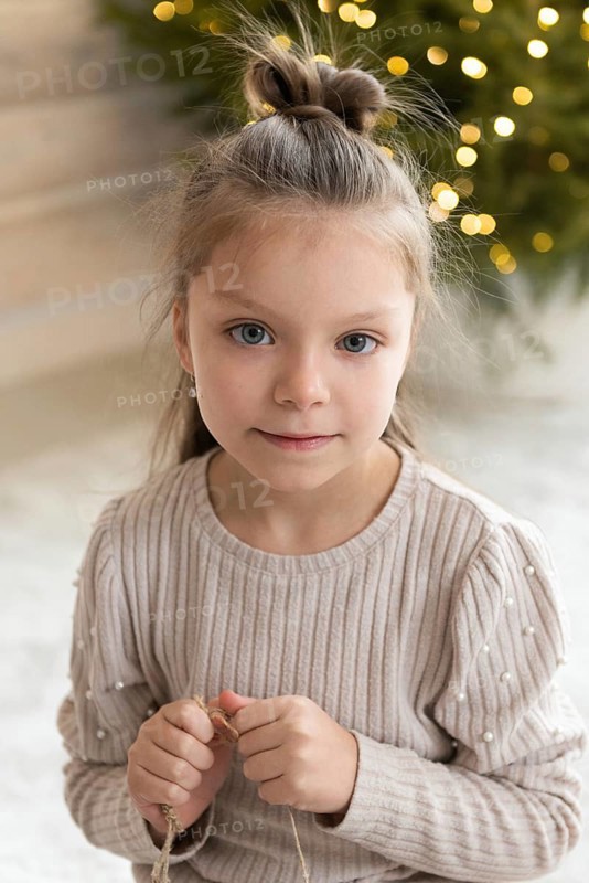 Close-up portrait of a young girl celebrating Christmas. She is looking directly at the camera with a subtle smile, dressed in a cozy sweater against a background softly lit by festive lights