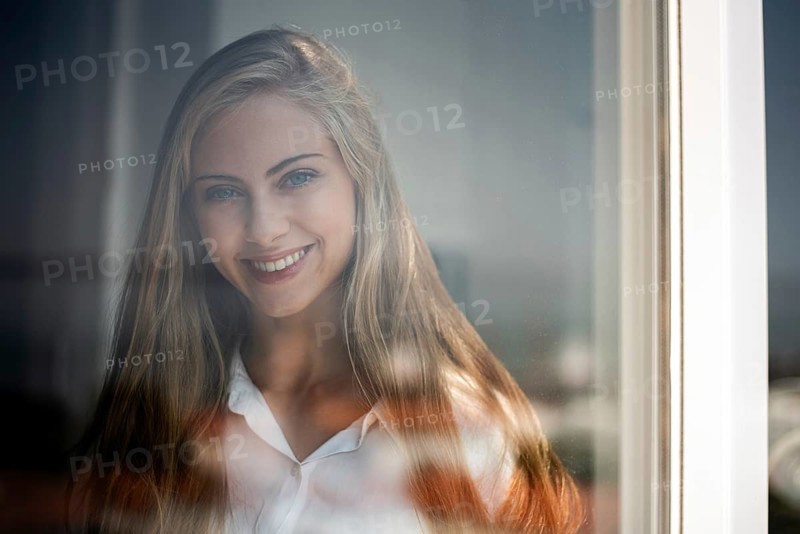 A young woman smiles gently through a glass window, captured in soft natural light. Her relaxed expression and casual attire create a serene and inviting atmosphere