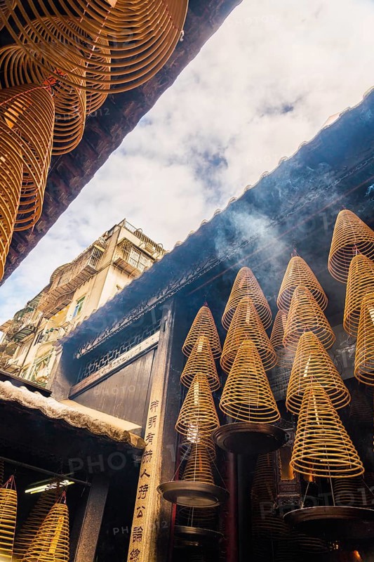 Spiral incense cones hang within the dimly lit interior of a Taoist temple in Macau, emitting fragrant smoke, against a backdrop of traditional architecture