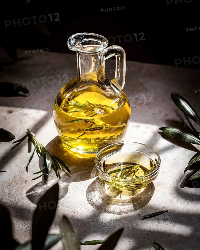A clear glass pitcher filled with golden olive oil, infused with fresh rosemary sprigs, sits elegantly on a rustic table. Sunlight casts dynamic shadows, enhancing its natural appeal