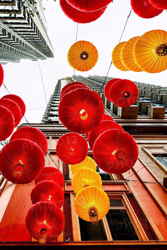 From below radiant red and yellow lanterns hang between skyscrapers in Hong Kong, celebrating Chinese New Year. The festive decorations create a vibrant canopy against the urban backdrop, symbolizing prosperity and good fortune