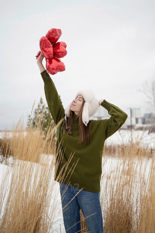 A young woman in a green sweater and white hat lifts red heart shaped balloons in a snowy park. The serene moment captures winter charm and romance