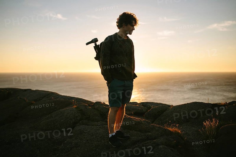 A young male photographer stands on the rocky terrain of Cape Spear Lighthouse, Newfoundland & Labrador, Canada, looking contemplatively at the sunset over the ocean while holding his camera
