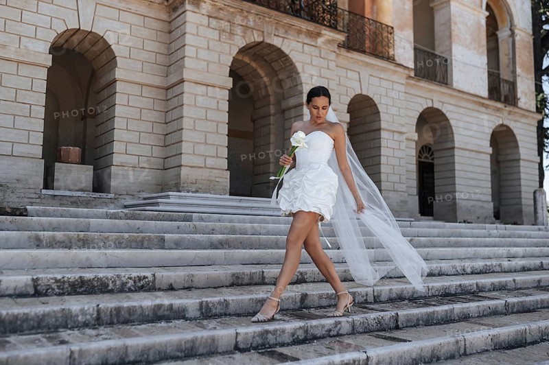 A beautiful bride in a white dress and veil gracefully descends the ancient stone steps of a historic villa in Treia, Macerata, Italy, holding a white flower