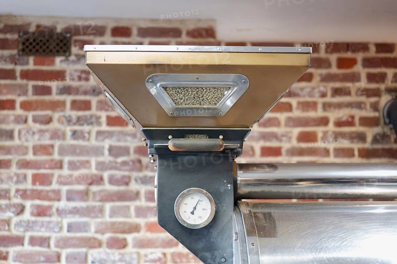 An industrial coffee roasting machine is showcased against a rustic brick wall. The machine holds green coffee beans, preparing for the precise roasting process
