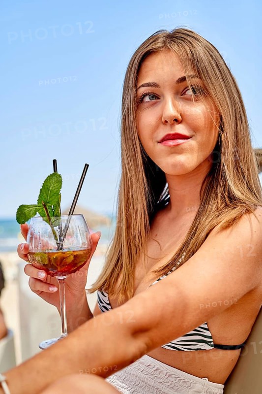 A young woman relaxes at the beach, enjoying a colorful cocktail while soaking up the sun. The waves gently lap at the shore in the background, enhancing the summer vibe
