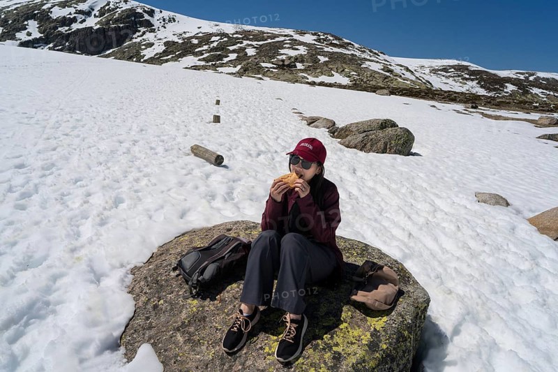 An Asian woman sits comfortably on a rocky surface, enjoying a meal amidst snow-covered peaks in the Sierra de Gredos, Spain. The clear sky adds to the serene ambiance