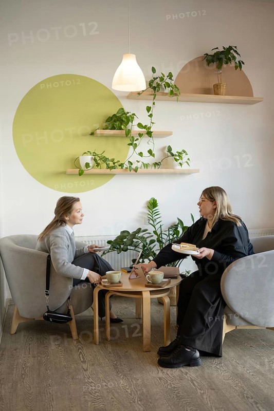 Two women enjoy a friendly chat over coffee in a cozy, stylish cafe with modern decor and lush greenery. Perfect setting for relaxation and meaningful conversation