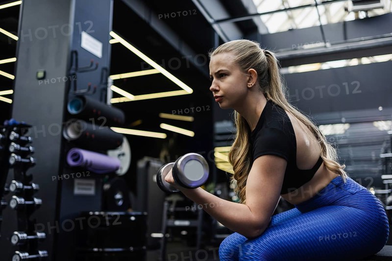 A focused blonde woman with blond hair performs bicep curls in a contemporary gym. She is wearing a black top and blue leggings, surrounded by fitness equipment and bright lighting