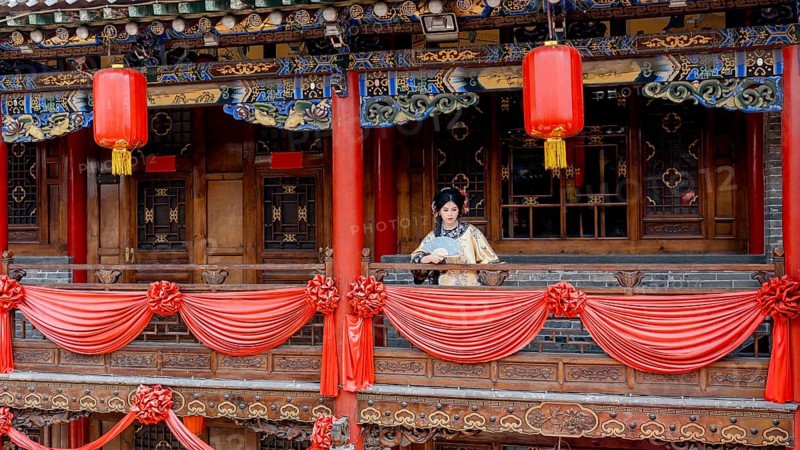 A woman in traditional Qing Dynasty attire stands on a beautifully decorated balcony in Pingyao, China. Red lanterns and intricate woodwork adorn the historical setting