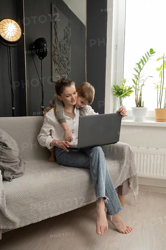 A mother working on a laptop at home with her young child lovingly embracing her. A cozy, modern interior with plants and warm lighting creates a comforting atmosphere