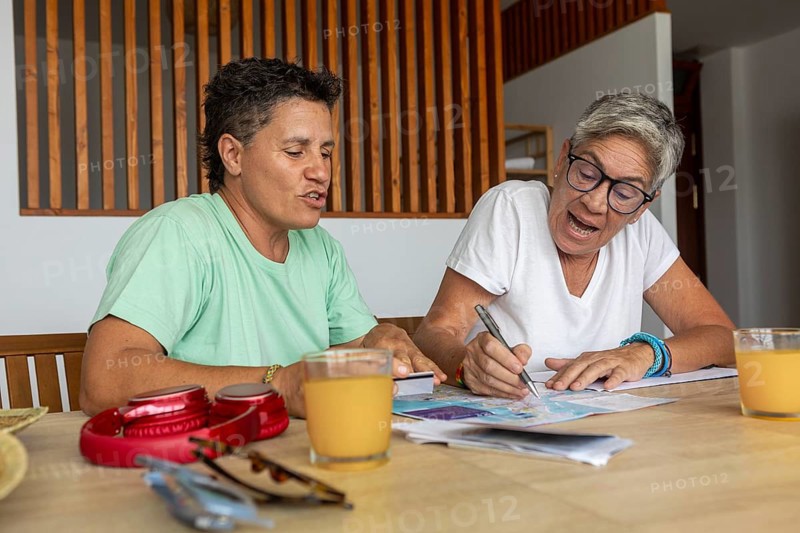 A lesbian couple is focused and active as they plan their summer holidays, marking details on travel brochures and discussing options excitedly over breakfast